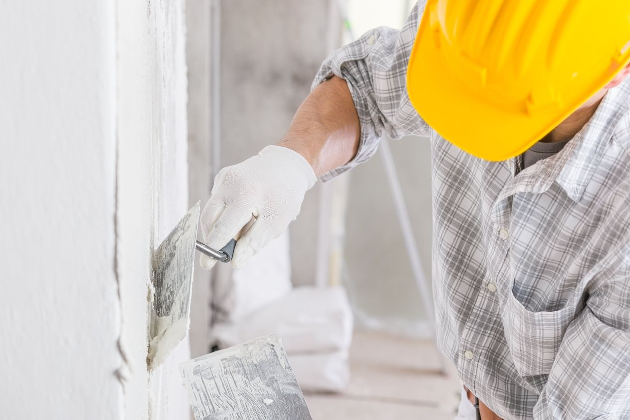 Ein hell gekleideter Handwerker mit gelben Helm verputzt eine weiße Wand